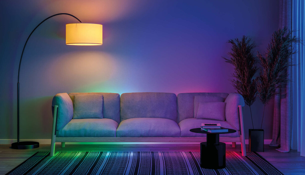 Licht als Designelement – Grün-blaupink beleuchtete Couch mit einer großen Lampe mit warmem Licht darüber.