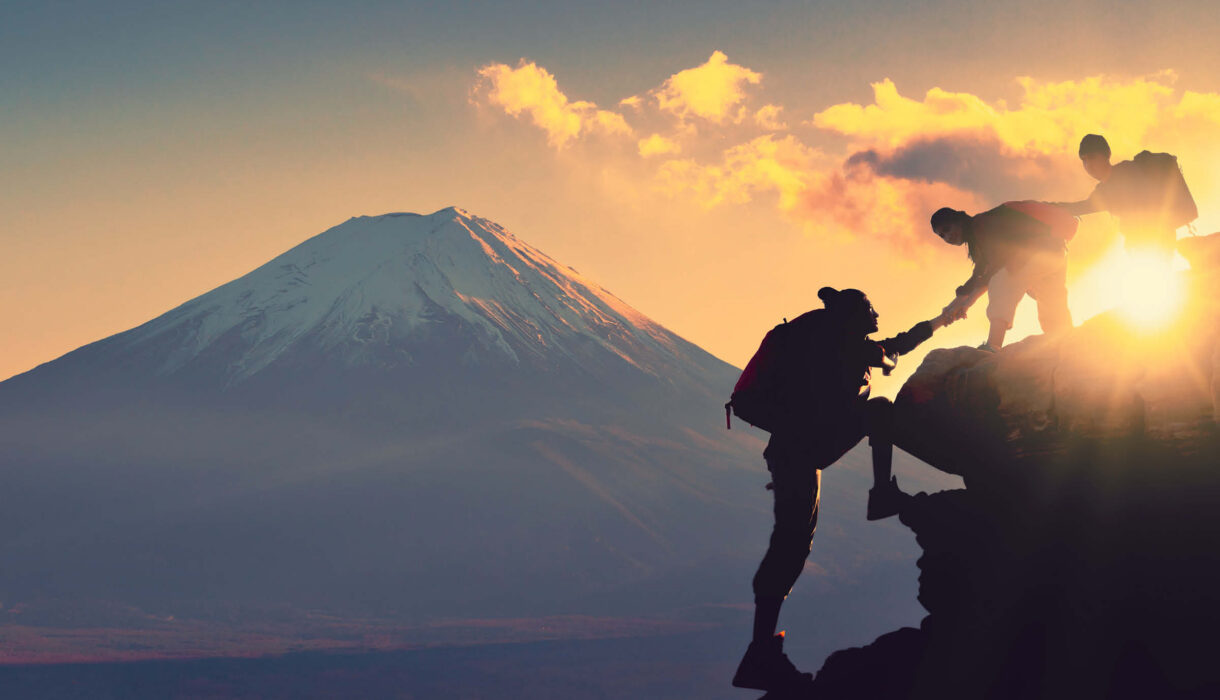 Bild von drei Bergsteigern, die sichgegenseitig nach oben untersützen. Im Hintergrund ist ein Sonnenuntergang zu sehen.