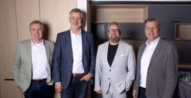 Vier Männer in Businesskleidung stehen nebeneinander in moderner Innenraumumgebung mit Holz- und Küchenelementen, Gruppenporträt von Geschäftsleuten bei Event oder Meeting