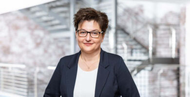 Professionelles Porträt von Astrid Strick von Geyso in modernem Unternehmensumfeld. Die Branchenexpertin trägt einen dunkelblauen Blazer und eine Brille und steht vor einer zeitgemäßen Architektur mit Treppenanlage