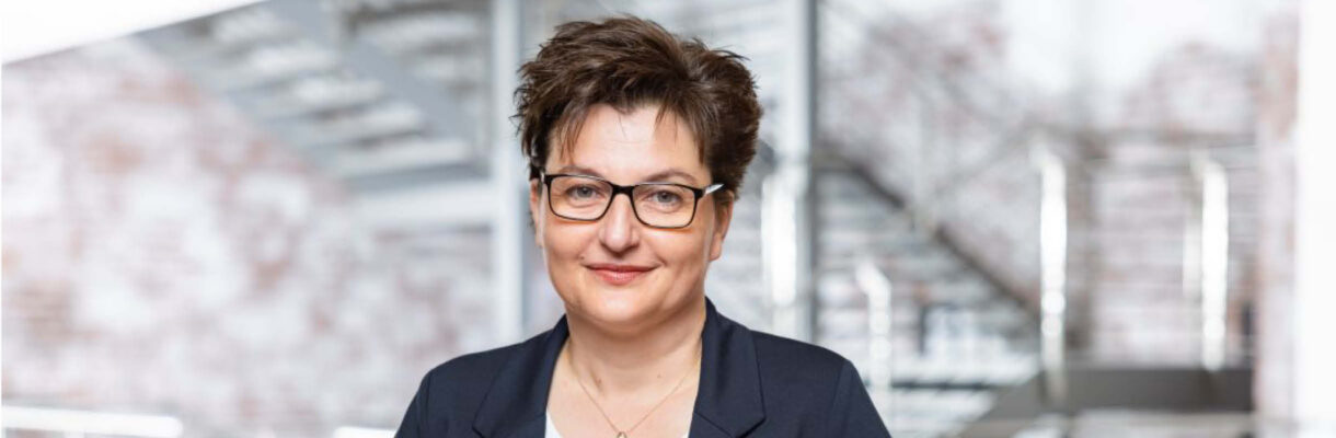 Professionelles Porträt von Astrid Strick von Geyso in modernem Unternehmensumfeld. Die Branchenexpertin trägt einen dunkelblauen Blazer und eine Brille und steht vor einer zeitgemäßen Architektur mit Treppenanlage