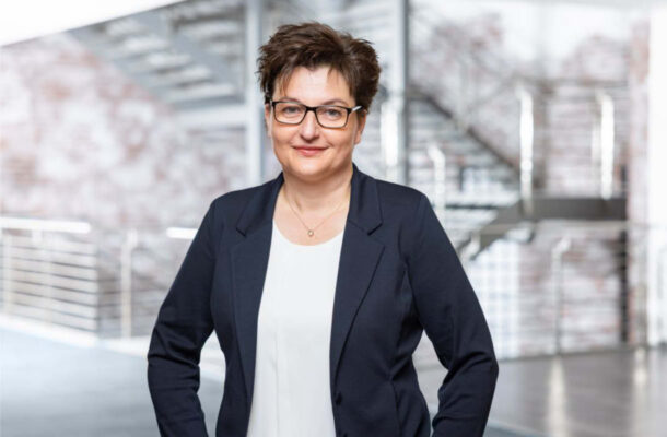 Professionelles Porträt von Astrid Strick von Geyso in modernem Unternehmensumfeld. Die Branchenexpertin trägt einen dunkelblauen Blazer und eine Brille und steht vor einer zeitgemäßen Architektur mit Treppenanlage