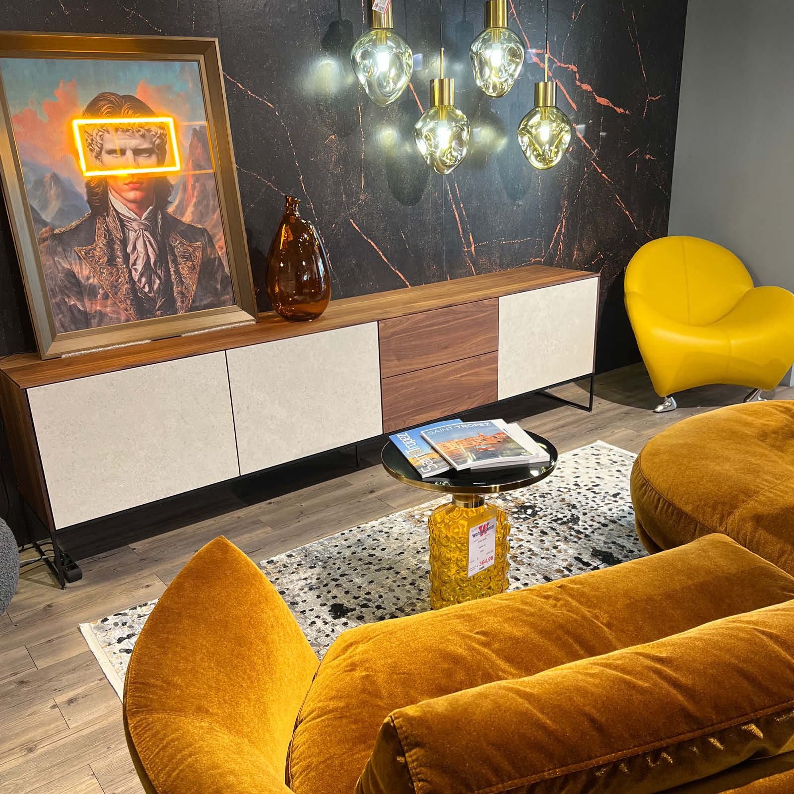 Moderner Wohnbereich im Möbelhaus mit Sideboard in Holzoptik, gelbem Sessel, Samtsofa, Designleuchten und Dekoration als Einrichtungsidee für offene Wohn- und Küchenbereiche.
