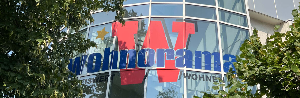 Außenansicht des Wohnorama Möbelhauses mit großer Glasfassade, blau-rotem Logo und grünen Bäumen – Standort für Möbel, Küchenmöbel und Inspiration rund um Küchenplanung und Wohnen.