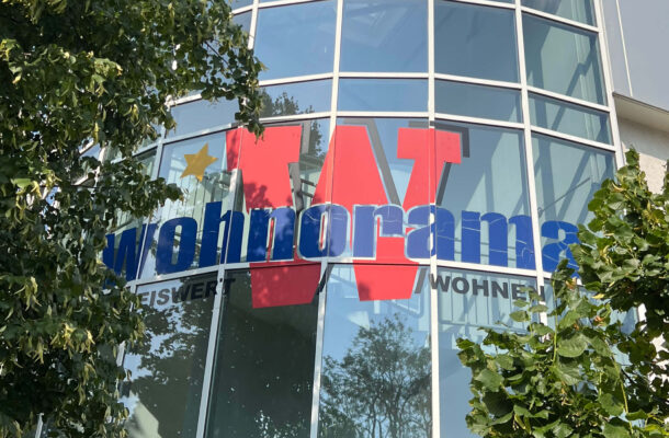 Außenansicht des Wohnorama Möbelhauses mit großer Glasfassade, blau-rotem Logo und grünen Bäumen – Standort für Möbel, Küchenmöbel und Inspiration rund um Küchenplanung und Wohnen.