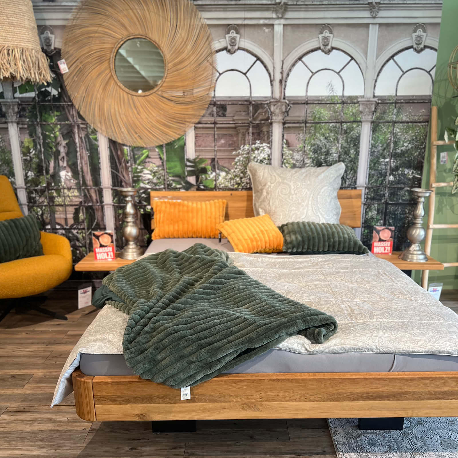 Stilvoll eingerichtetes Schlafzimmer im Möbelhaus mit Holzbett, grüner Tagesdecke, Dekokissen, Wandmotiv und natürlichen Materialien als Inspiration für Wohnen, Möbel und angrenzende Küchenbereiche.