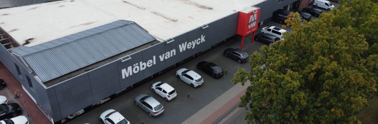 Luftaufnahme des Möbelhauses Möbel van Weyck mit großer grauer Fassade, Kundenparkplätzen und weitläufigem Flachdach – Standort für Möbel, Küchenstudio, individuelle Küchenplanung und Einrichtung.
