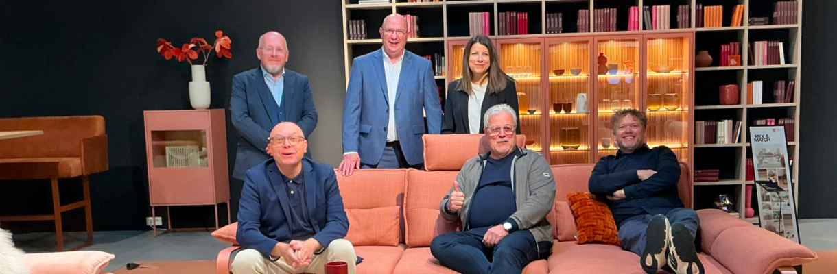 Messebesucher auf einem korallfarbenen Sofa vor einer beleuchteten Regalwand auf der Möbelmesse.