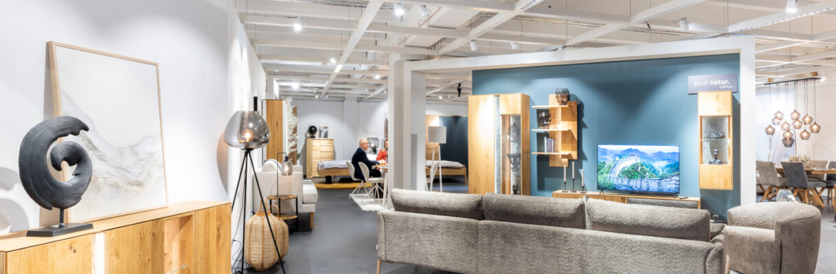 Möbelausstellung mit grauem Sofa, Sessel und Holzmöbeln auf einer großzügigen Ausstellungsfläche.