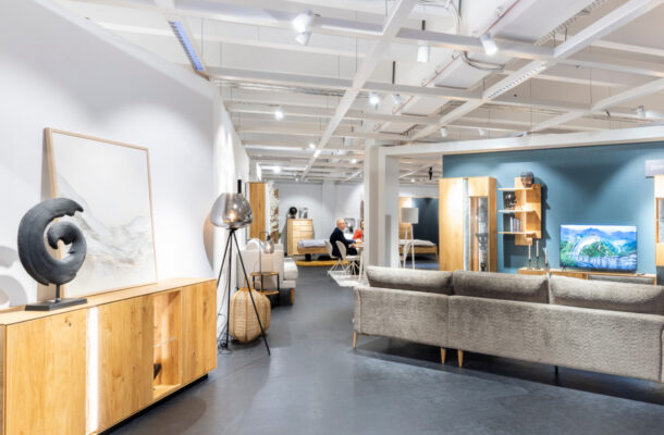 Möbelausstellung mit grauem Sofa, Sessel und Holzmöbeln auf einer großzügigen Ausstellungsfläche.